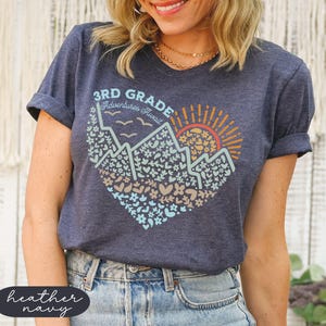 K&ouml;nnte beinhalten: Marineblaues Heather-T-Shirt mit herzf&ouml;rmiger Grafik, die Berge, eine Sonne und den Text "3rd Grade Adventures Await" zeigt. Das Design enth&auml;lt Blumen- und Herzmotive in verschiedenen Farben.