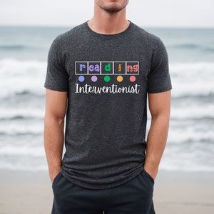 Op de afbeelding: Een donkergrijs T-shirt met het woord "lezen" gespeld in kleurrijke vierkanten met een cirkel in elk vierkant. De tekst "Interventionist" staat onder het woord "lezen".