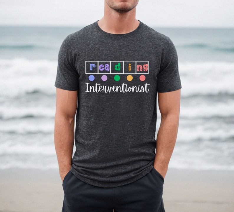 Op de afbeelding: Een donkergrijs T-shirt met het woord "lezen" gespeld in kleurrijke vierkanten met een cirkel in elk vierkant. De tekst "Interventionist" staat onder het woord "lezen".