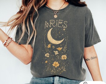 May include: A gray t-shirt with a graphic design of a crescent moon, stars, and flowers. The text "Aries" is printed in gold lettering above the moon.