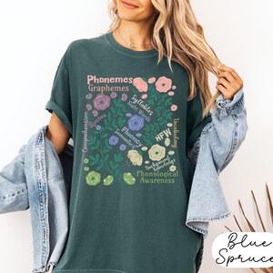 May include: A teal-colored t-shirt with a floral design containing educational terms like "Phonemes" and "Graphemes." The design includes pink, purple, and yellow flowers. The words "Blue Spruce" are written in a cursive font at the bottom right.