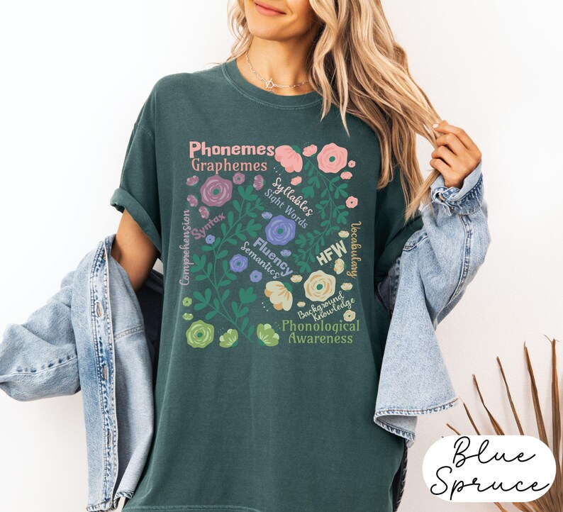 May include: A teal-colored t-shirt with a floral design containing educational terms like "Phonemes" and "Graphemes." The design includes pink, purple, and yellow flowers. The words "Blue Spruce" are written in a cursive font at the bottom right.