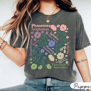 May include: Dark gray t-shirt with a floral design featuring educational terms like "Phonemes," "Graphemes," and "Fluency." The design includes various flowers in shades of pink, purple, and cream, with green leaves. The shirt has a relaxed fit.