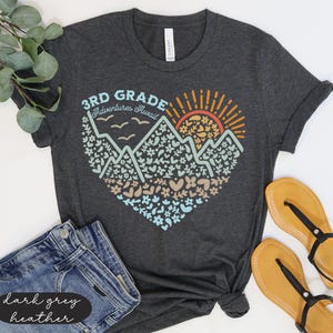 K&ouml;nnte beinhalten: Dunkelgraues Heather-T-Shirt mit einem herzf&ouml;rmigen Aufdruck mit Bergen, Blumen und einer aufgehenden Sonne. Der Text lautet "3rd Grade Adventures Await". Das Shirt wird mit blauen Jeansshorts und beigen Sandalen kombiniert.