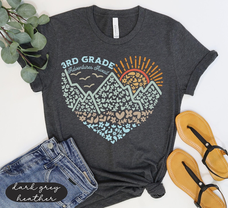K&ouml;nnte beinhalten: Dunkelgraues Heather-T-Shirt mit einem herzf&ouml;rmigen Aufdruck mit Bergen, Blumen und einer aufgehenden Sonne. Der Text lautet "3rd Grade Adventures Await". Das Shirt wird mit blauen Jeansshorts und beigen Sandalen kombiniert.
