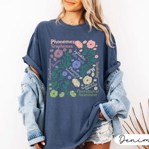 May include: A navy blue t-shirt with a floral design featuring the words "Phonemes, Graphemes, Syllables, Sight Words, Fluency, Semantics, HFW, Phonological Awareness". The shirt has a relaxed fit and is made of a soft material.