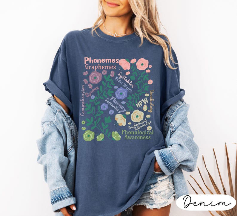 May include: A navy blue t-shirt with a floral design featuring the words "Phonemes, Graphemes, Syllables, Sight Words, Fluency, Semantics, HFW, Phonological Awareness". The shirt has a relaxed fit and is made of a soft material.