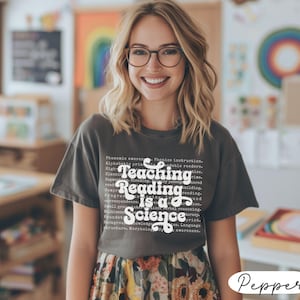 May include: A dark gray t-shirt with the text "Teaching Reading is a Science" in a white, stylized font. The shirt also features smaller text related to reading and education. The person is wearing glasses and a floral skirt.