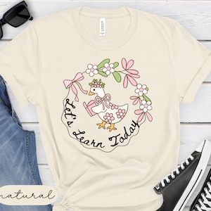 May include: Cream-colored t-shirt featuring a cartoon duck wearing a floral crown and holding a book, surrounded by a floral wreath. The text "Let's Learn Today" is written around the duck. The shirt is made of a soft material.