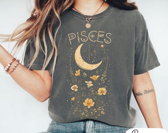 Pode incluir: Uma camiseta cinza com um design gráfico de uma lua crescente, estrelas e flores. O texto "Pisces" está impresso em letras douradas acima da lua.