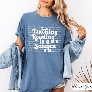 May include: A blue jean-colored t-shirt with the text "Teaching Reading is a Science" in white script. The shirt has rolled-up sleeves and is worn with a denim jacket. The text includes words related to reading and education.