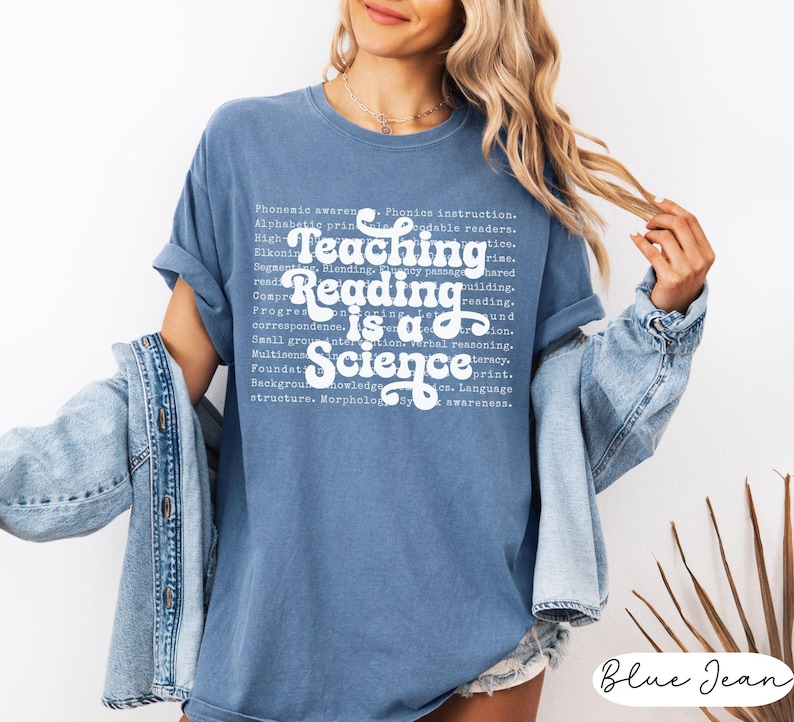 May include: A blue jean-colored t-shirt with the text "Teaching Reading is a Science" in white script. The shirt has rolled-up sleeves and is worn with a denim jacket. The text includes words related to reading and education.