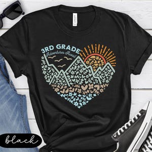 K&ouml;nnte beinhalten: Schwarzes T-Shirt mit einem herzf&ouml;rmigen Design mit Bergen, Blumen und einer aufgehenden Sonne. Der Text "3rd Grade Adventures Await" ist &uuml;ber der Grafik aufgedruckt. Das T-Shirt wird mit blauen Jeans und schwarzen Turnschuhen getragen.