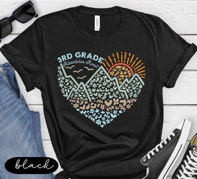 K&ouml;nnte beinhalten: Schwarzes T-Shirt mit einem herzf&ouml;rmigen Design mit Bergen, Blumen und einer aufgehenden Sonne. Der Text "3rd Grade Adventures Await" ist &uuml;ber der Grafik aufgedruckt. Das T-Shirt wird mit blauen Jeans und schwarzen Turnschuhen getragen.