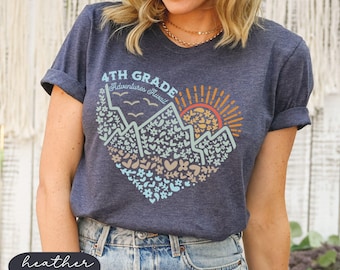 Puede incluir: Camiseta azul marino con un gráfico en forma de corazón que presenta montañas, un sol y el texto "4th Grade Adventures Await". El diseño incorpora elementos florales y geométricos en tonos azules, naranjas y beige. La camiseta es de material suave.