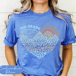 K&ouml;nnte beinhalten: Columbia-blaues T-Shirt mit einem grafischen Design. Das Design zeigt Berge, eine Sonne und den Text "3rd Grade Adventures Await". Die Grafik ist in Blau-, Orange- und Beiget&ouml;nen gehalten.