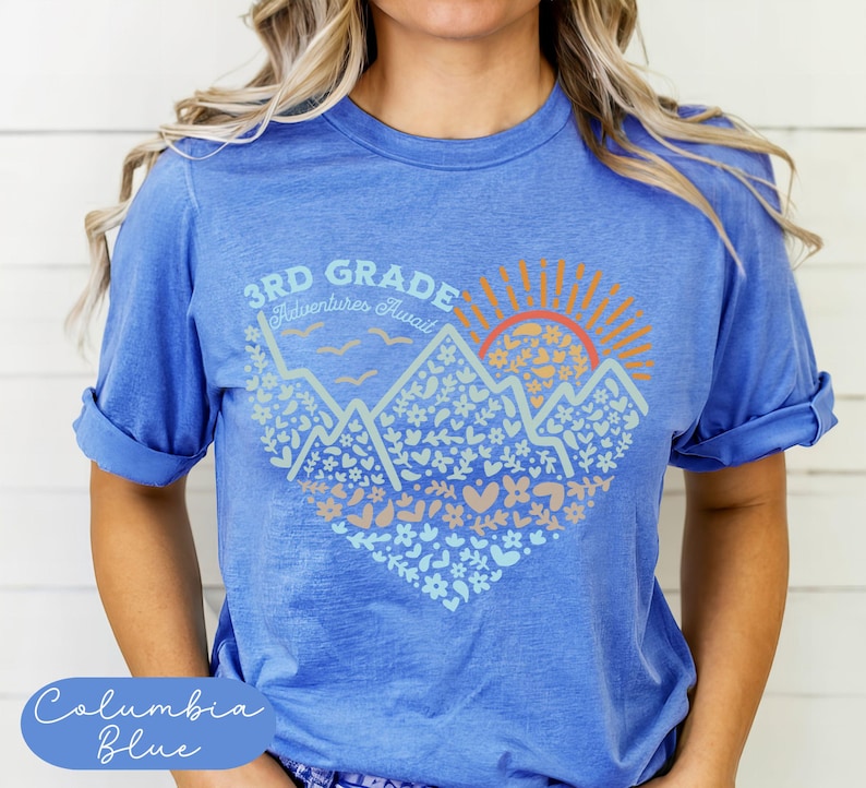 K&ouml;nnte beinhalten: Columbia-blaues T-Shirt mit einem grafischen Design. Das Design zeigt Berge, eine Sonne und den Text "3rd Grade Adventures Await". Die Grafik ist in Blau-, Orange- und Beiget&ouml;nen gehalten.