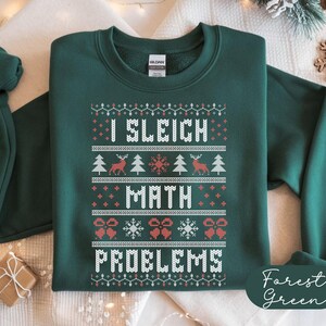 以下が含まれることがあります： 「I Sleigh Math Problems」の白文字がプリントされたフォレストグリーンのスウェットシャツ。トナカイ、雪の結晶、クリスマスツリーがフェスティバル模様でデザインされています。スウェットシャツはGildanブランドです。