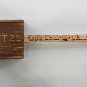 Op de afbeelding: Een houten sigarenkistgitaar met een liniaal voor het toetsbord. De liniaal is gelabeld "Hardy Groth Blacksmith Supply". De gitaar heeft een gouden logo met de tekst "RPETIZE Gourmet Grains".