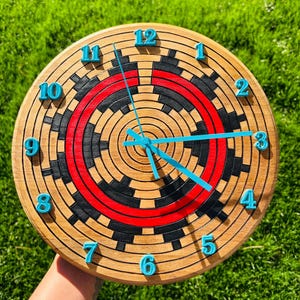 May include: A circular wooden clock with a distinctive design. The clock face has a concentric pattern of black, red, and natural wood tones. Teal blue numbers and hands provide contrast. The clock is held against a background of green grass.