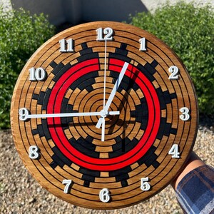 May include: A round wooden clock with a brown finish, featuring white numerals and hands. The clock face has a unique design with black and red concentric circles. The clock is approximately 12 inches in diameter.