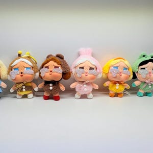 Puede incluir: Seis muñecas de peluche con ojos grandes y detalles de pedrería. Cada muñeca está vestida con un atuendo diferente, que incluye un vestido azul, un disfraz de abeja, un disfraz de oso, un atuendo rosa, un atuendo amarillo y un disfraz de rana.