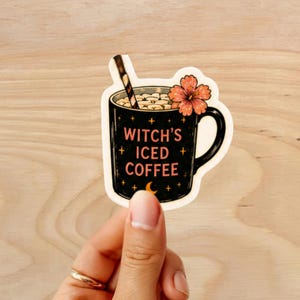Puede incluir: Una pegatina con una taza negra con la inscripci&oacute;n "Witch's Iced Coffee". La taza tiene un asa, una pajita y una flor rosa. La pegatina est&aacute; sujeta por una mano.