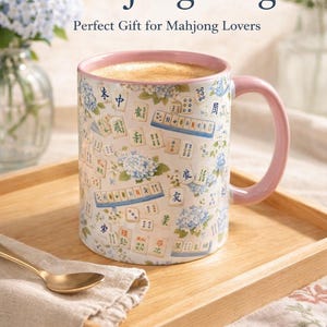 May include: A white ceramic mug with a pink handle and rim, decorated with Mahjong tiles and blue floral accents. The mug is filled with a light brown liquid and sits on a wooden tray. Text on the mug reads "Beautiful Mahjong Mug".