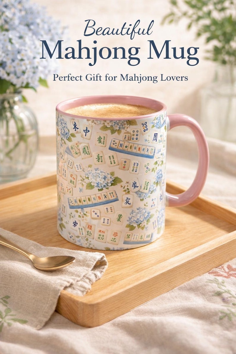 May include: A white ceramic mug with a pink handle and rim, decorated with Mahjong tiles and blue floral accents. The mug is filled with a light brown liquid and sits on a wooden tray. Text on the mug reads "Beautiful Mahjong Mug".
