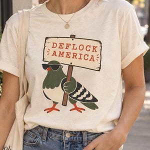 May include: A cream-colored short-sleeve t-shirt featuring a graphic of a pigeon wearing black sunglasses and holding a wooden sign that reads DEFLOCK AMERICA in red letters. The pigeon has green, black, and white feathers with orange feet.