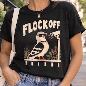 May include: A black short-sleeve t-shirt featuring a graphic print of a white pigeon wearing black sunglasses. The bird stands near a security camera, with the text FLOCKOFF printed in large cream-colored letters above the illustration.