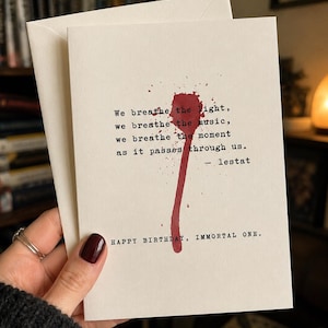 May include: A cream-colored greeting card with a red ink splatter design resembling a blood drip. The text reads: We breathe the light, we breathe the music, we breathe the moment as it passes through us. - lestat. Below is: HAPPY BIRTHDAY, IMMORTAL ONE.