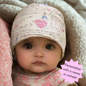 May include: A beige baby beanie featuring a pink heart with the name "emily" printed on it. The hat has a floral and script design with a Peter Rabbit illustration. The image includes the text "Personalize with a name or initials!"