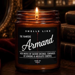 May include: A lit candle in an amber glass jar with a black label. The label reads: SMELLS LIKE THE VAMPIRE Armand WITH NOTES OF SACRED INCENSE, CENTURIES OF LONGING & ABSOLUTE CONTROL MADE IN PARIS. A thin red vertical line runs through the text.