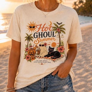 Op de afbeelding: Beige t-shirt met een kleurrijke afbeelding van een spook, zwarte kat en pompoen op een strand. De tekst luidt "Hot Ghoul Summer" met extra tekst: "Sun, Sand, Spirits, Good Vibes & Spooky Times."