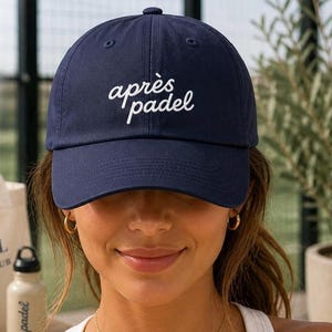 May include: Navy blue baseball cap with the text "après padel" in white script. The cap has a curved brim and a fabric strap closure. A classic, casual accessory for sports enthusiasts.
