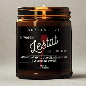 May include: A brown glass jar candle with a lit wick. The label reads "Smells Like The Vampire Lestat de Lioncourt" with scent notes. The candle is made in New Orleans.