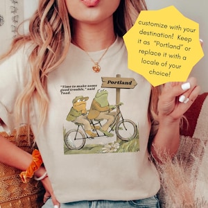 Pode incluir: Camiseta creme com uma ilustração lúdica de dois sapos em uma bicicleta tandem, indo em direção a uma placa de "Portland". O texto diz: "Time to make some good trouble, said Toad."