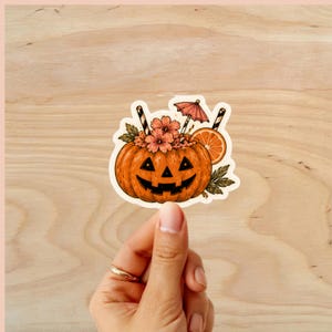 Puede incluir: Una pegatina con tem&aacute;tica de Halloween que presenta una calabaza naranja con una cara tallada, llena de flores y una rodaja de naranja. La calabaza tiene dos pajitas a rayas y un peque&ntilde;o paraguas rosa. La pegatina est&aacute; sujeta por una mano.
