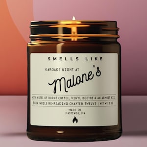 May include: A brown glass jar candle with a white label that reads: SMELLS LIKE KAROAKE NIGHT AT Malone's WITH NOTES OF BURNT COFFEE, VINYL BOOTHS & AN ALMOST KISS BURN WHILE RE-READING CHAPTER TWELVE | NET WT. 9 OZ MADE IN HASTINGS, MA.