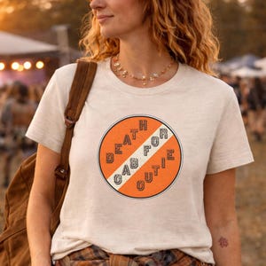 May include: A beige t-shirt with a circular orange graphic that reads "DEATH FOR CUTIE" in a vintage style. The text is in white, with a white stripe through the center of the circle. The shirt is short-sleeved.