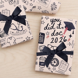 May include: Gift-wrapped presents with navy blue bows. The wrapping paper features medical-themed illustrations and text that reads "You did it, doc 2026." Other illustrations include stethoscopes, scrubs, and coffee cups.