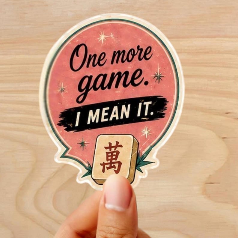 Może przedstawiać: Okrągła naklejka z r&oacute;żowym i białym wzorem. Naklejka zawiera czarny tekst "One more game. I MEAN IT.". Ręka trzyma naklejkę. Na dole znajduje się płytka z chińskim znakiem.