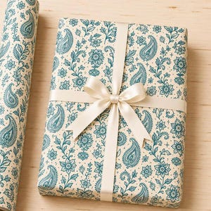 May include: A rectangular gift box wrapped in cream-colored paper with a teal paisley and floral pattern. The box is tied with a smooth, cream-colored satin ribbon featuring a centered bow. A matching roll of patterned wrapping paper sits nearby.