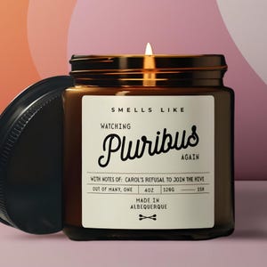 May include: A lit candle in a brown glass jar with a black lid. The label reads "Smells Like Watching Pluribus Again" with additional text about the scent and origin. The candle is 4 oz and burns for 15 hours.