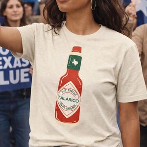 Pode incluir: Camiseta bege com gola redonda e um gráfico vermelho de uma garrafa de molho picante. A garrafa tem uma tampa verde e o texto diz "Flip Tables Talarico Texas Original". A camiseta é de manga curta.