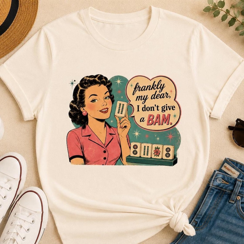 May include: Off-white t-shirt with a vintage-style illustration of a woman holding a card. The graphic includes the text "frankly my dear, I don't give a BAM." Below the woman are illustrated mahjong tiles. The woman is wearing a pink shirt.