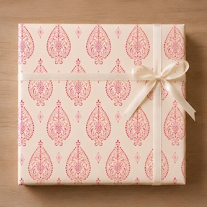 May include: A square gift box wrapped in cream-colored paper featuring a repeating red botanical leaf pattern. The box is tied with a smooth, cream-colored satin ribbon that is knotted into a bow on the right side of the package.