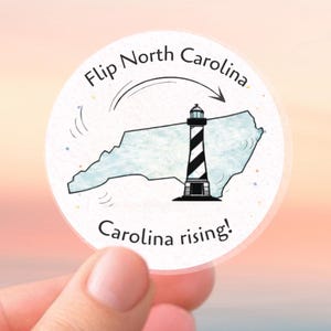Puede incluir: Una pegatina redonda con un mapa de Carolina del Norte y un faro con rayas blancas y negras. El texto "Flip North Carolina" y "Carolina rising!" están escritos en la pegatina.