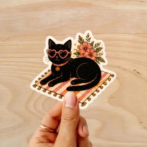 Puede incluir: Una pegatina con un gato negro con gafas de sol en forma de coraz&oacute;n y un collar de calabaza, tumbado en una alfombra a rayas. Hay flores detr&aacute;s del gato. La pegatina est&aacute; sobre una superficie de madera.